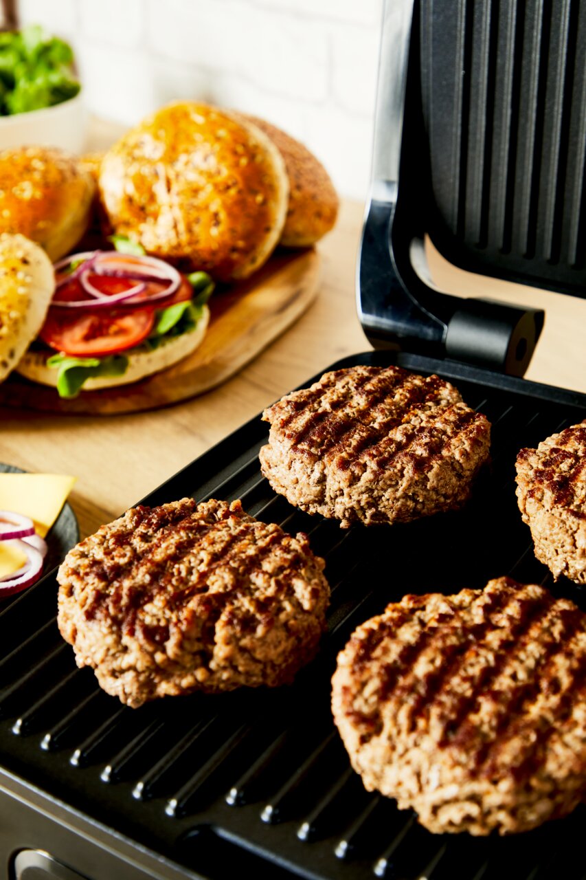 Kontaktgrill mit gegrillten Burger-Patties, im Hintergrund frische Burgerbrötchen und Zutaten, seitliche Perspektive