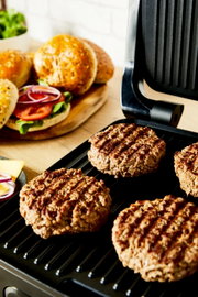 Contactgrill met gegrilde hamburgers, op de achtergrond verse hamburgerbroodjes en ingrediënten, zijaanzicht