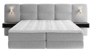 Frontale Ansicht eines grauen Boxspringbetts mit gepolstertem Kopfteil, integrierten Nachttischen und Wandlampen.