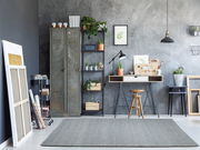 Moderner Raum mit grauem Handweb Teppich im Vordergrund, umgeben von industriellen und natürlichen Elementen wie Metallspind, Pflanzen und Holzmöbeln.