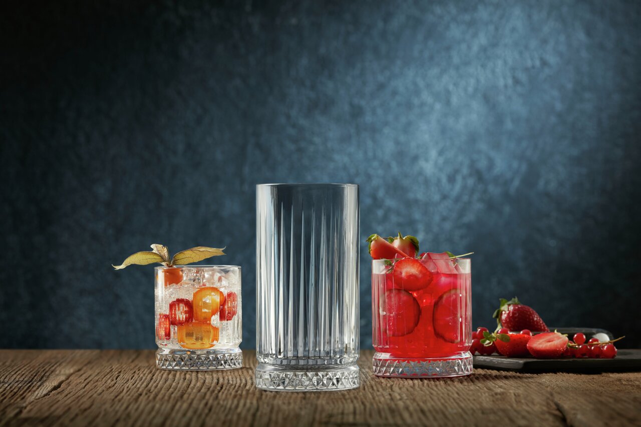 Drei Longdrinkbecher aus dem Elysia 4er Set auf einem Holztisch, frontal fotografiert. Zwei Becher sind mit Getränken und Früchten gefüllt, der mittlere Becher ist leer.