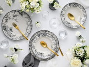 Draufsicht auf elegantes Tischgedeck mit schwarz-weiß floral gemusterten Porzellantellern, goldfarbenem Besteck, klaren Gläsern und weißen Blumenarrangements auf hellem Hintergrund.