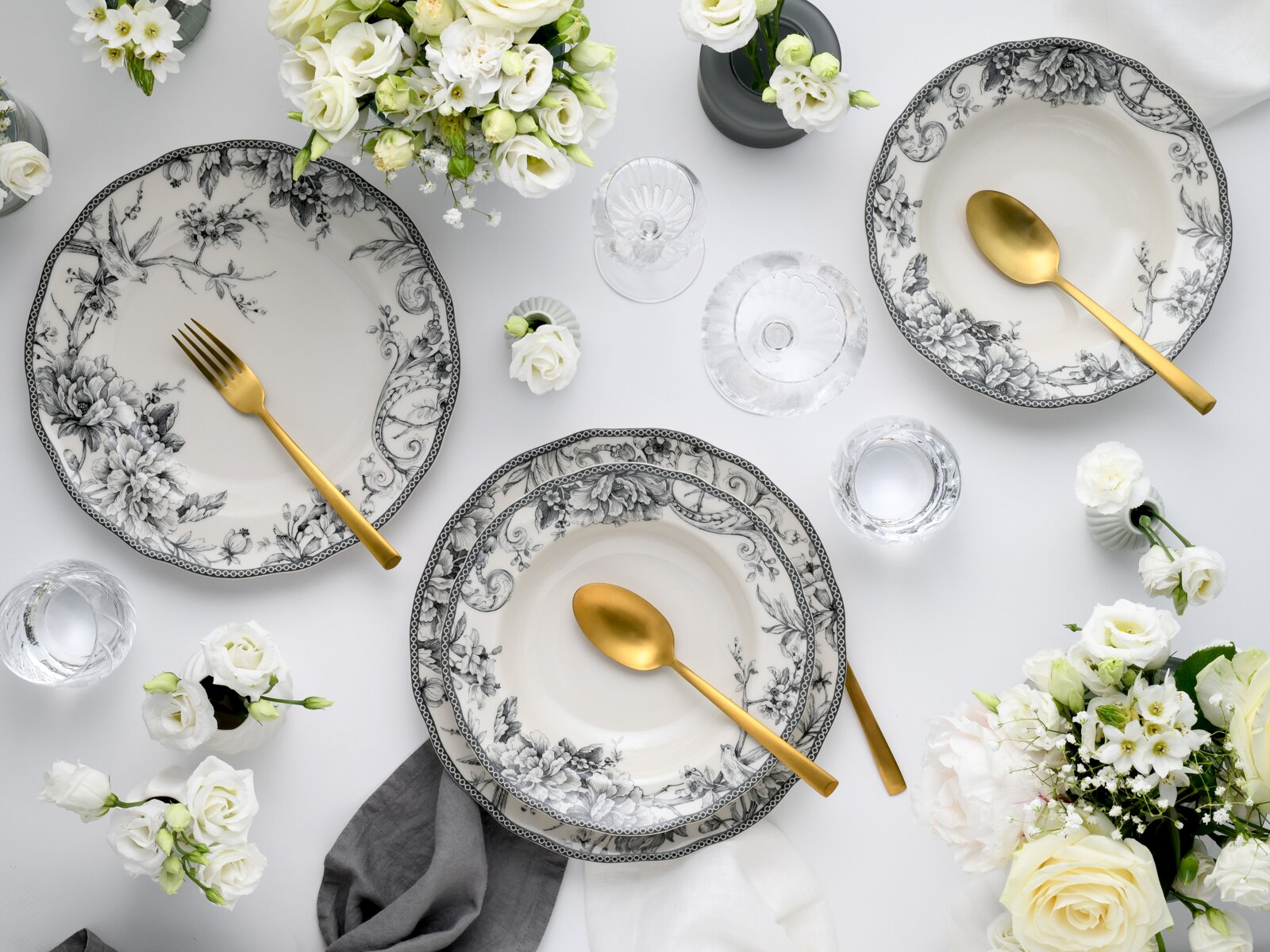Draufsicht auf elegantes Tischgedeck mit schwarz-weiß floral gemusterten Porzellantellern, goldfarbenem Besteck, klaren Gläsern und weißen Blumenarrangements auf hellem Hintergrund.