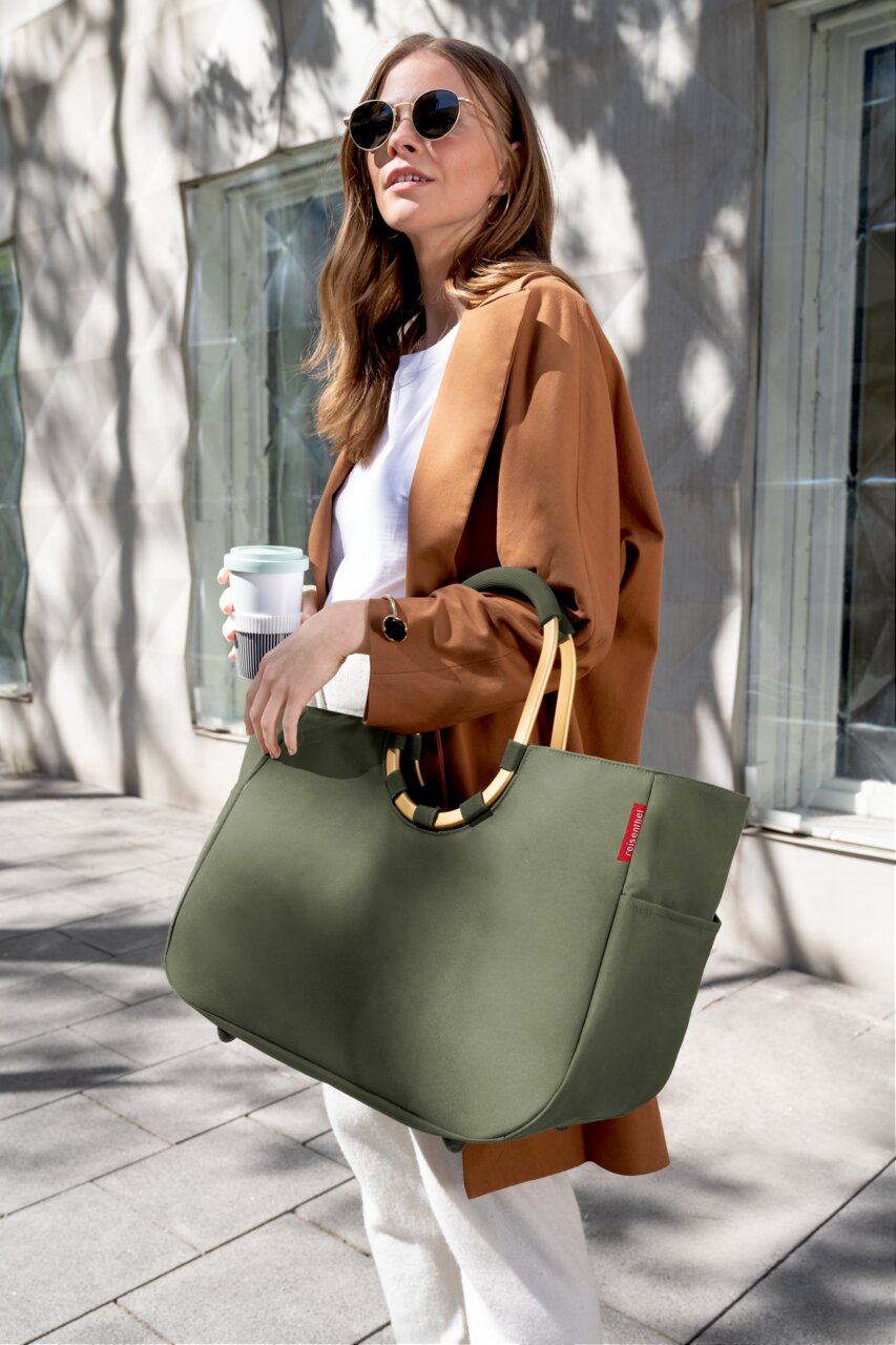 Reisenthel Loopshopper L Urban Forest Gold Frau in Sonnenbrille und braunem Mantel trägt eine große, olivgrüne Tasche mit runden Griffen und genießt einen sonnigen Tag in der Stadt. Perspektive: seitlich von vorne.