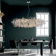 Moderner Kronleuchter RICICLE mit funkelnden Kristallen, hängend über einem stilvollen Esstisch in einem elegant eingerichteten Raum, seitliche Perspektive.