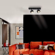 Schwarzer Deckenstrahler mit zwei verstellbaren Spots auf rechteckiger Schiene, eingeschaltet, in modernem Wohnzimmer über orangefarbener Couch; Perspektive: leicht von unten im Raum nach oben zur Decke fotografiert.