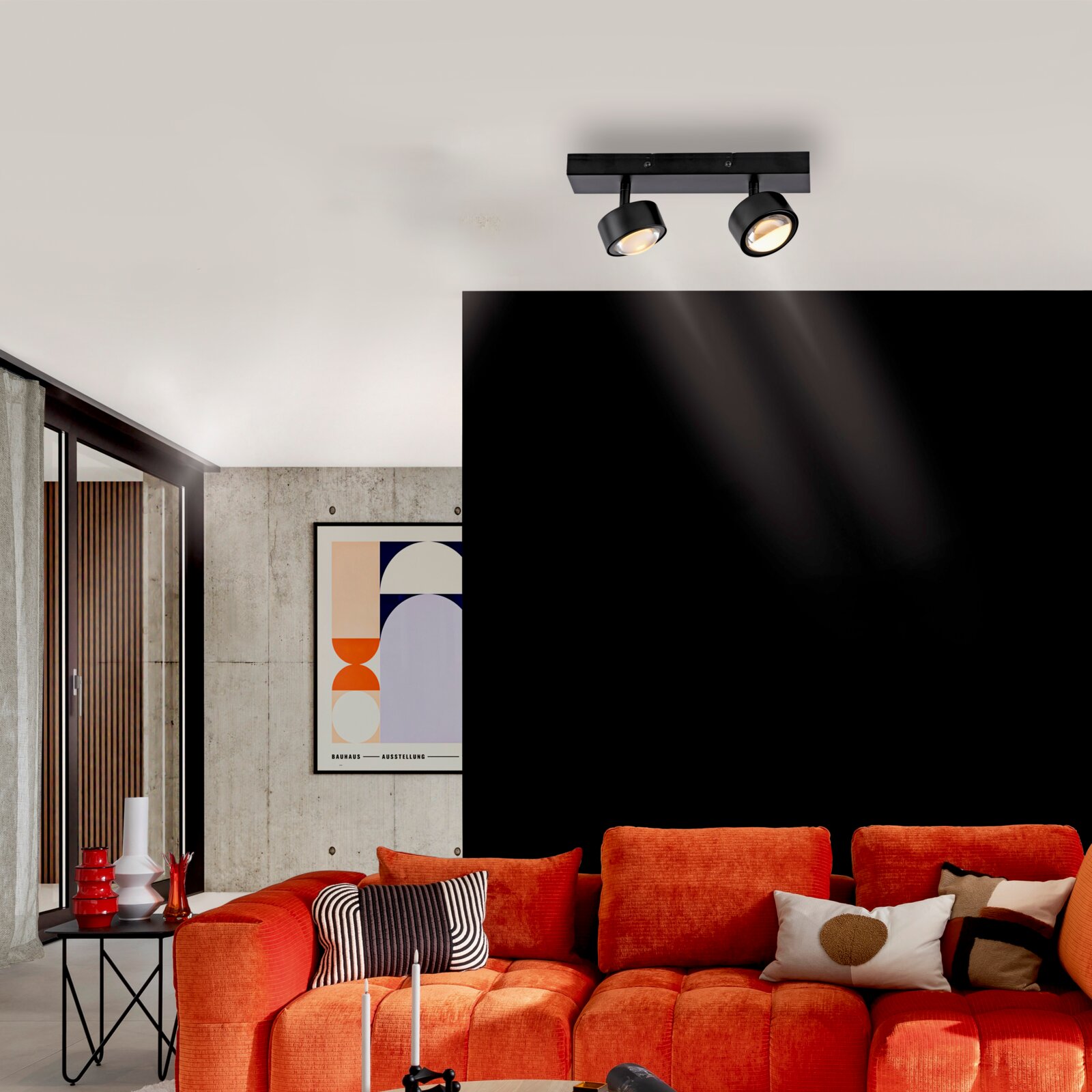 Schwarzer Deckenstrahler mit zwei verstellbaren Spots auf rechteckiger Schiene, eingeschaltet, in modernem Wohnzimmer über orangefarbener Couch Perspektive: leicht von unten im Raum nach oben zur Decke fotografiert.