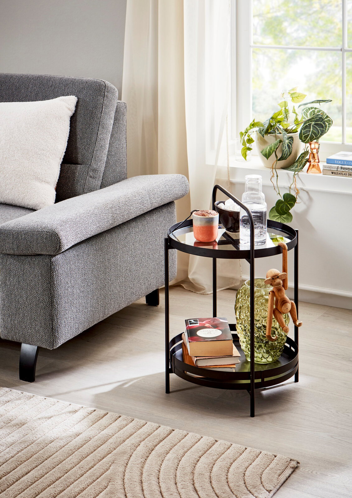 MID.YOU Couchtisch SMALL Schwarzer, runder Servierwagen mit zwei Ablagen und Griff, neben grauem Sessel am Fenster, mit Glasflasche, Tasse, Büchern und Deko – Perspektive: leicht schräg von vorne auf Augenhöhe.