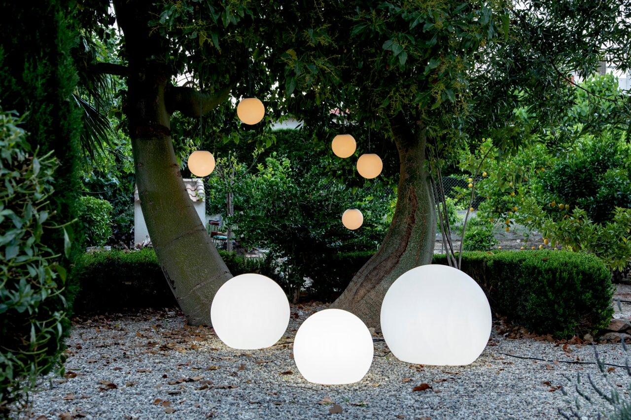 BULY 50-bolvormige lampen in een tuin, op de grond en hangend aan bomen, bij schemering gezien vanuit het vooraanzicht.