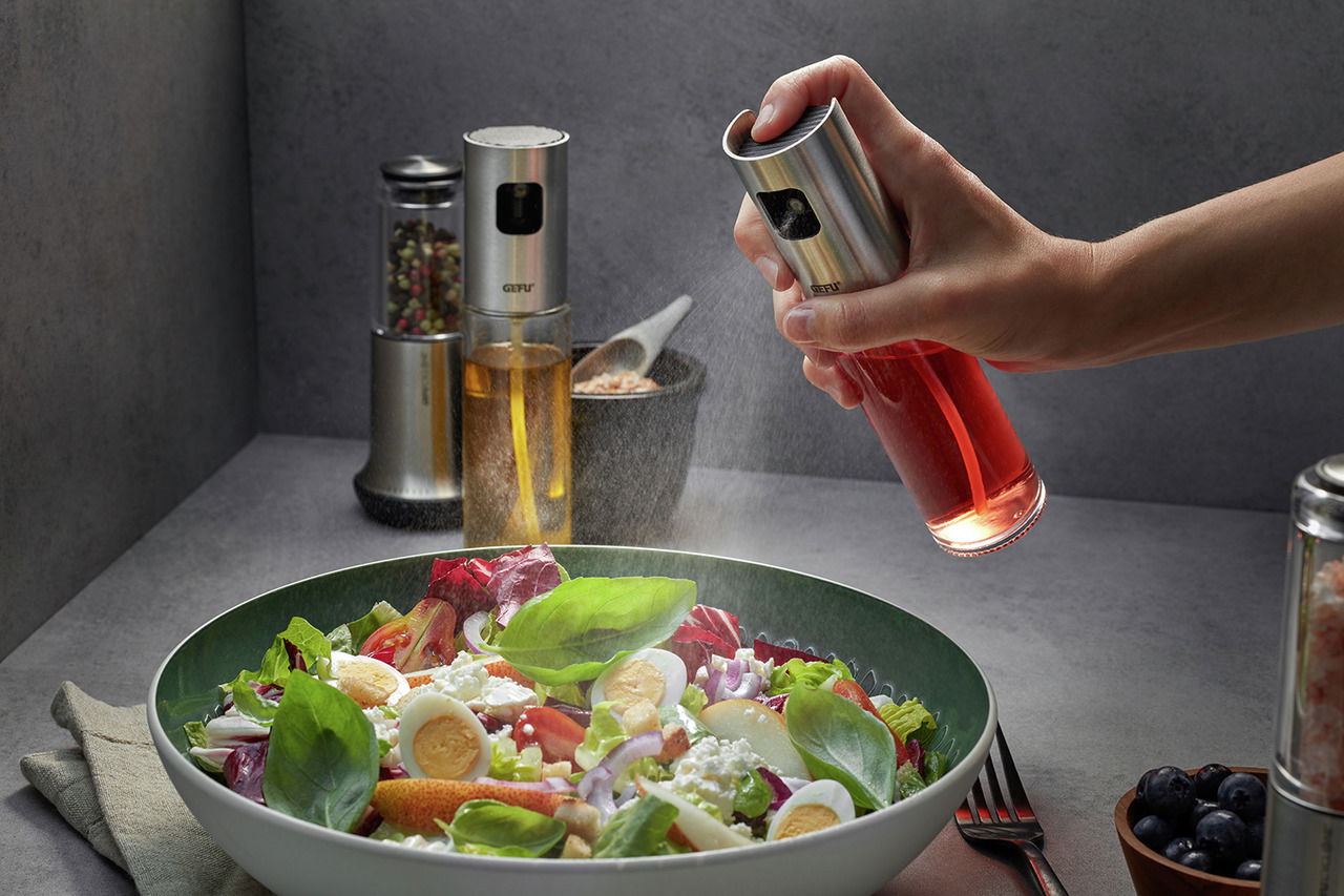 Essig-Öl-Sprüher NEVA in der Hand, der aus der Perspektive von oben auf einen frischen Salat sprüht.