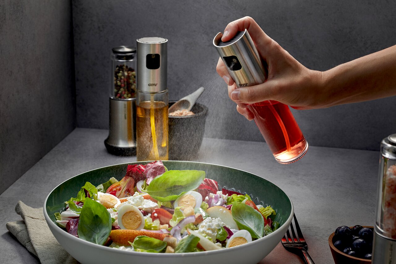 Essig-Öl-Sprüher NEVA in der Hand, der aus der Perspektive von oben auf einen frischen Salat sprüht.