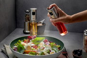 Essig-Öl-Sprüher NEVA in der Hand, der aus der Perspektive von oben auf einen frischen Salat sprüht.