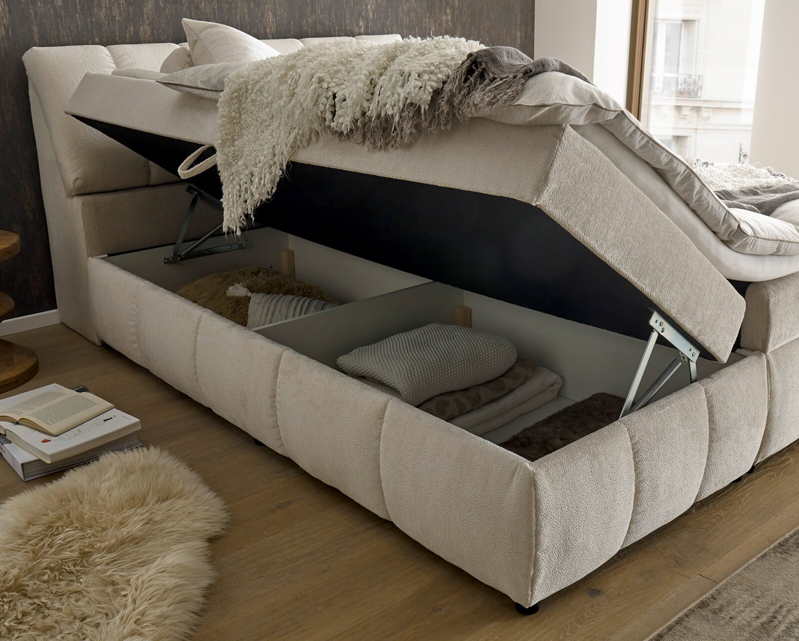 Boxspringbett mit geöffnetem Bettkasten, seitliche Perspektive, zeigt Stauraum mit Decken und Kissen.