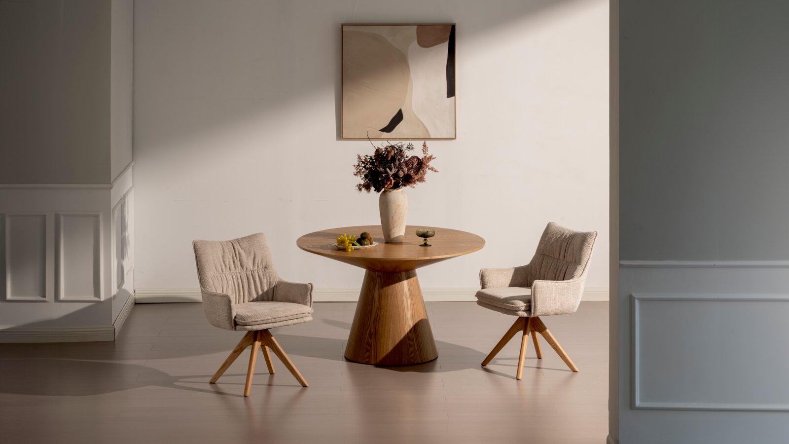Ronde houten eettafel met conische voet, gefotografeerd frontaal op ooghoogte, met twee beige gestoffeerde draaistoelen met houten poten in een minimalistische ruimte