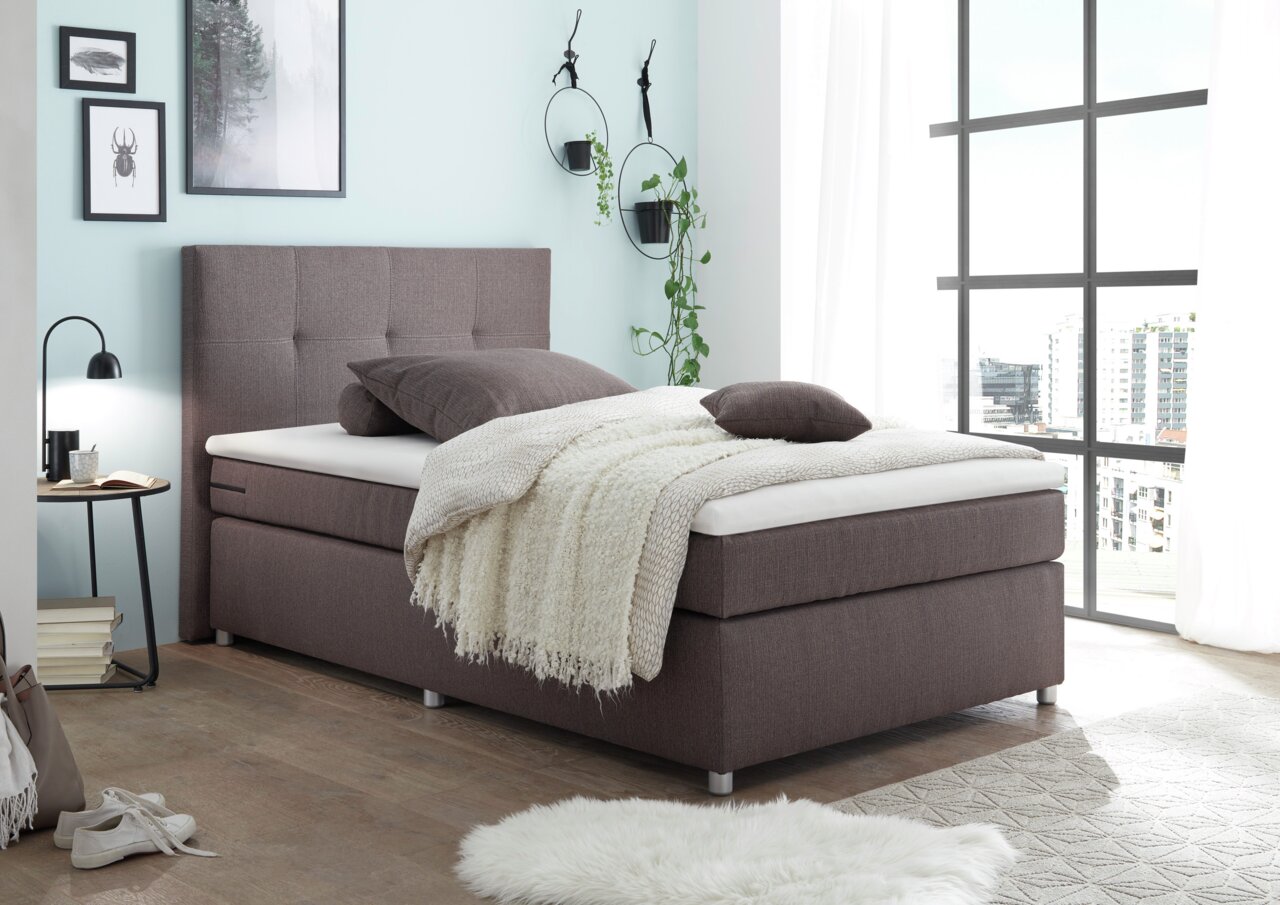 Esposa Boxspringbed MANOLO Boxspringbett in modernem Schlafzimmer, seitliche Perspektive, mit braunem Stoffbezug, dekoriert mit Kissen und Decke, umgeben von stilvollen Wanddekorationen und großen Fenstern.