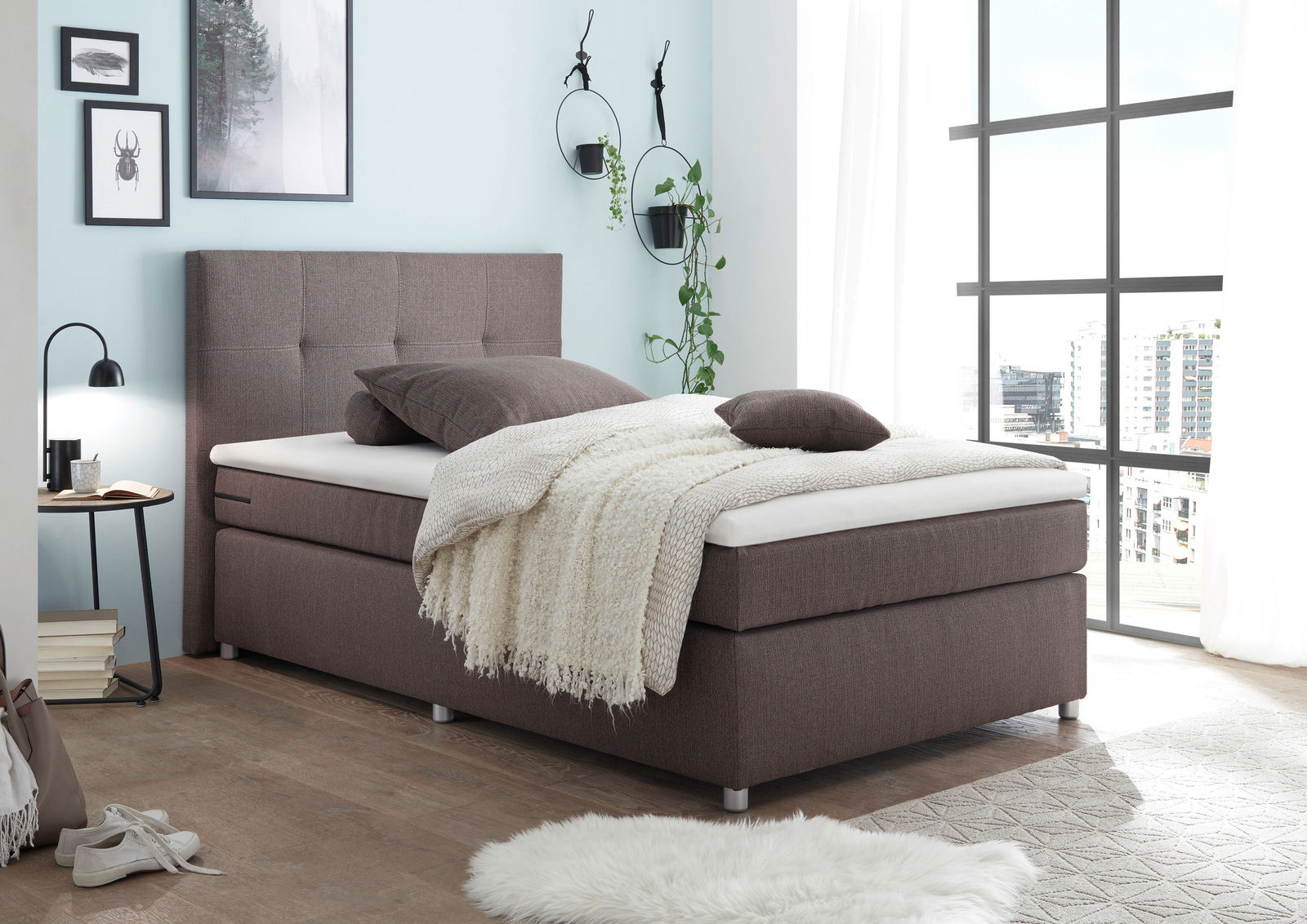 Esposa Boxspringbett MANOLO Boxspringbett in modernem Schlafzimmer, seitliche Perspektive, mit braunem Stoffbezug, dekoriert mit Kissen und Decke, umgeben von stilvollen Wanddekorationen und großen Fenstern.