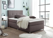 Boxspringbett in modernem Schlafzimmer, seitliche Perspektive, mit braunem Stoffbezug, dekoriert mit Kissen und Decke, umgeben von stilvollen Wanddekorationen und großen Fenstern.