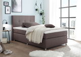 Boxspringbett in modernem Schlafzimmer, seitliche Perspektive, mit braunem Stoffbezug, dekoriert mit Kissen und Decke, umgeben von stilvollen Wanddekorationen und großen Fenstern.