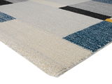 Nahaufnahme eines rechteckigen Teppichs mit geometrischem Muster in Blau-, Grau- und Beigetönen, perspektivisch von der Seite aufgenommen.