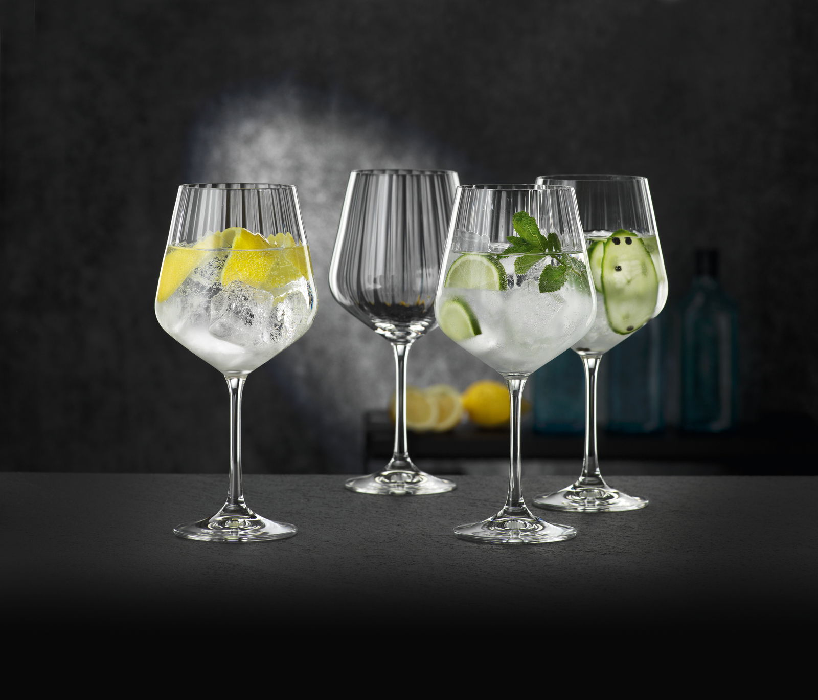 Vier elegante Gin Tonic Gläser, gefüllt mit Eis und verschiedenen Garnituren, auf einer dunklen Oberfläche vor einem stilvollen Hintergrund. Perspektive: seitlich.