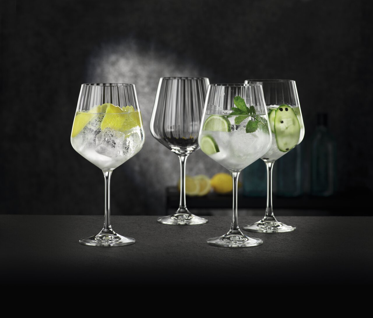 Vier elegante gin-tonicglazen, gevuld met ijs en diverse garneringen, op een donker oppervlak tegen een stijlvolle achtergrond. Perspectief: zijaanzicht.