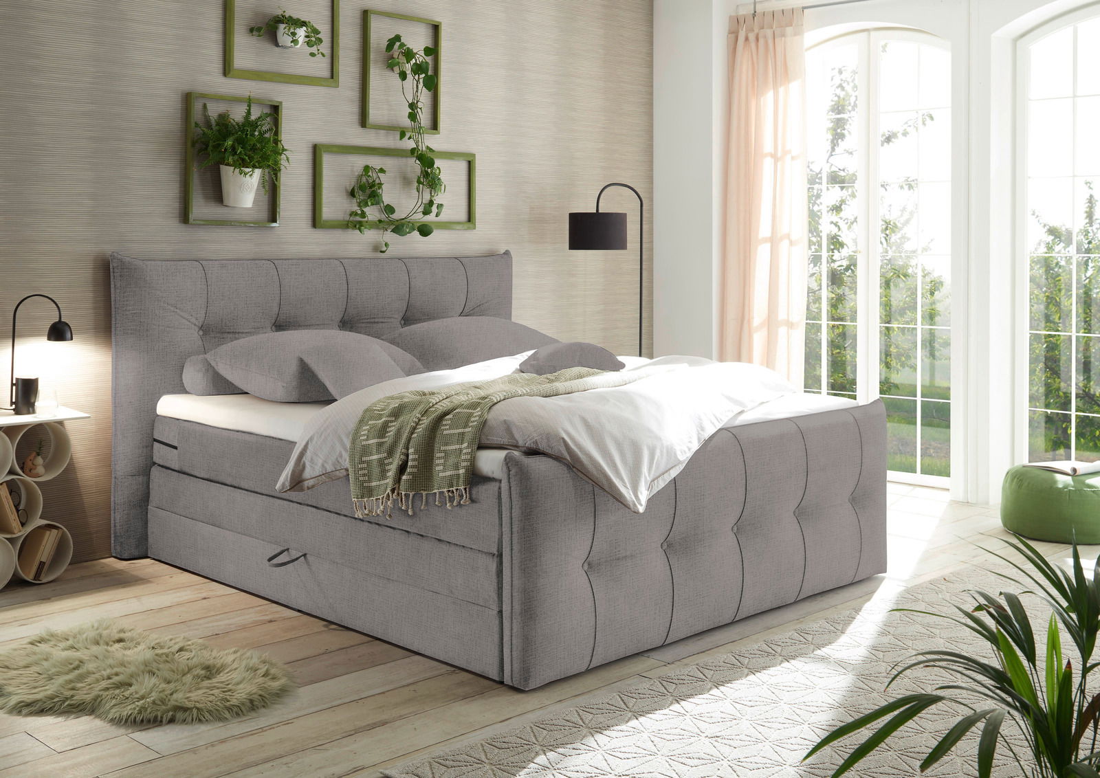 Gemütliches Boxspringbett in einem hellen Schlafzimmer mit großen Fenstern, Pflanzen und moderner Dekoration, seitliche Perspektive.