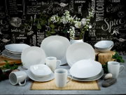 Weißes Porzellan-Geschirrset mit Tellern, Schalen und Bechern auf dekorativem Tisch-Arrangement vor Tafelwand mit Schriftzügen, frontal aus leicht erhöhter Perspektive fotografiert.