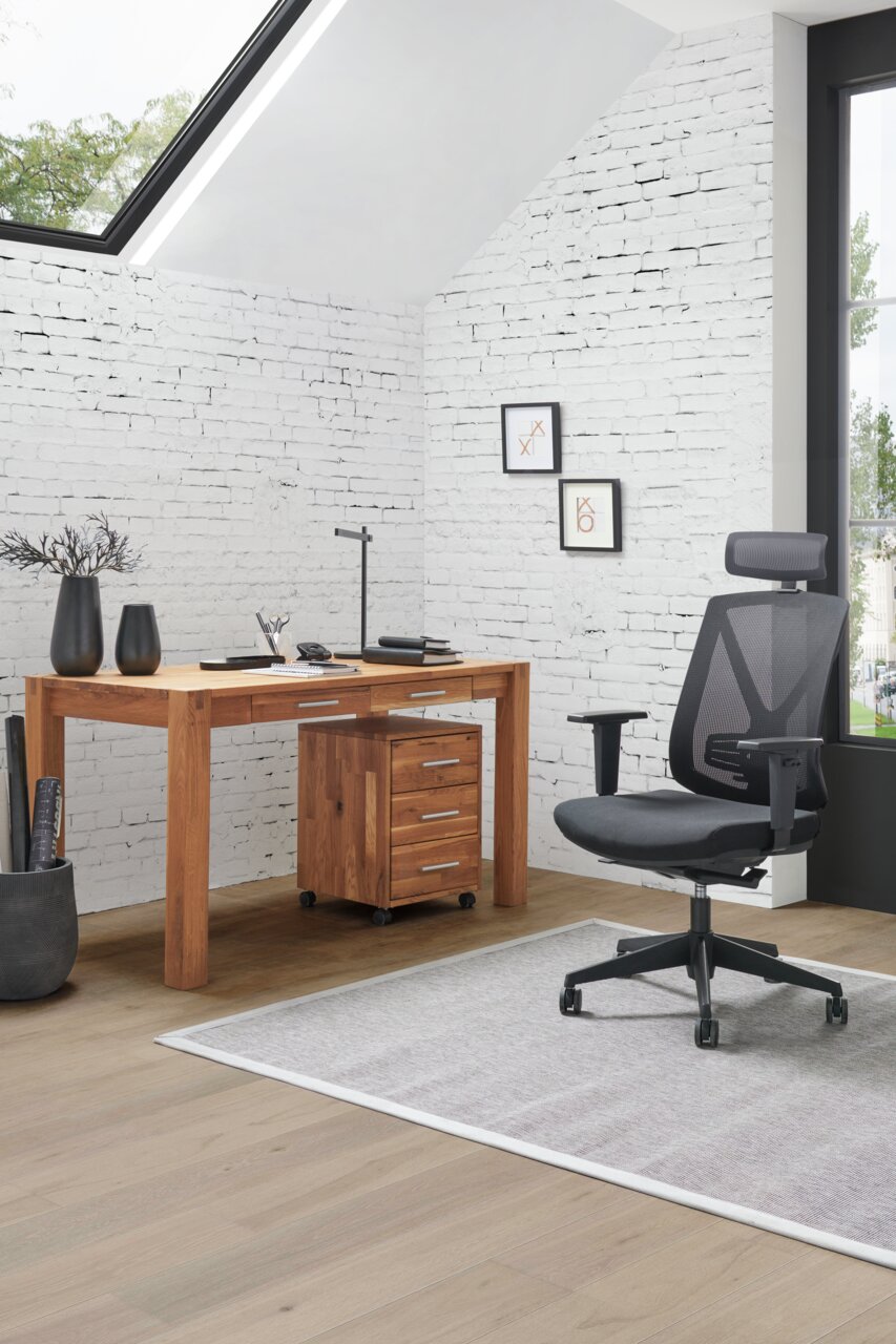 Moderner Schreibtisch aus Holz mit Schubladen, seitliche Perspektive, in einem hellen Raum mit weißer Backsteinwand und großem Fenster.