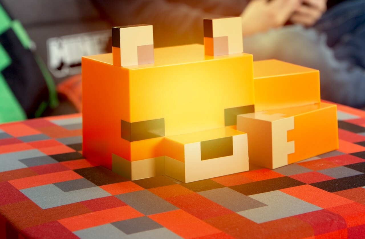 Orangefarbene, blockige Fuchs-Figur im Pixel-/Voxel-Design mit weißen Ohren und Schnauze auf rot-grau kariertem Tisch; Perspektive: leicht von vorne links in Augenhöhe aufgenommen.