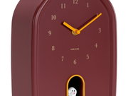 Moderne Wanduhr in Rot mit goldenen Zeigern und Zahlen, seitliche Perspektive, mit kleinem Fach und dekorativer Figur.