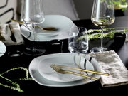 Modernes Tischgedeck mit weißen Tellern mit schwarzer und goldener Linien-Dekoration, goldfarbenem Besteck und Weingläsern auf schwarzem Tisch, fotografiert aus schräger Draufsicht.