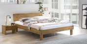 Massives Holzbett mit breitem Kopfteil in modernem, hellem Schlafzimmer, schräg von vorne in leichter Seitenansicht fotografiert.