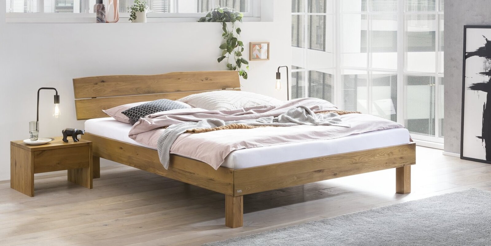 Massives Holzbett mit breitem Kopfteil in modernem, hellem Schlafzimmer, schräg von vorne in leichter Seitenansicht fotografiert.