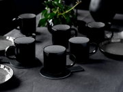 Mehrere schwarze Keramik-Kaffeetassen mit hell gesprenkelter Innenseite auf einem dunkel gedeckten Tisch, teils auf Untersetzern, fotografiert aus leicht erhöhter schräger Perspektive (3/4-Ansicht)