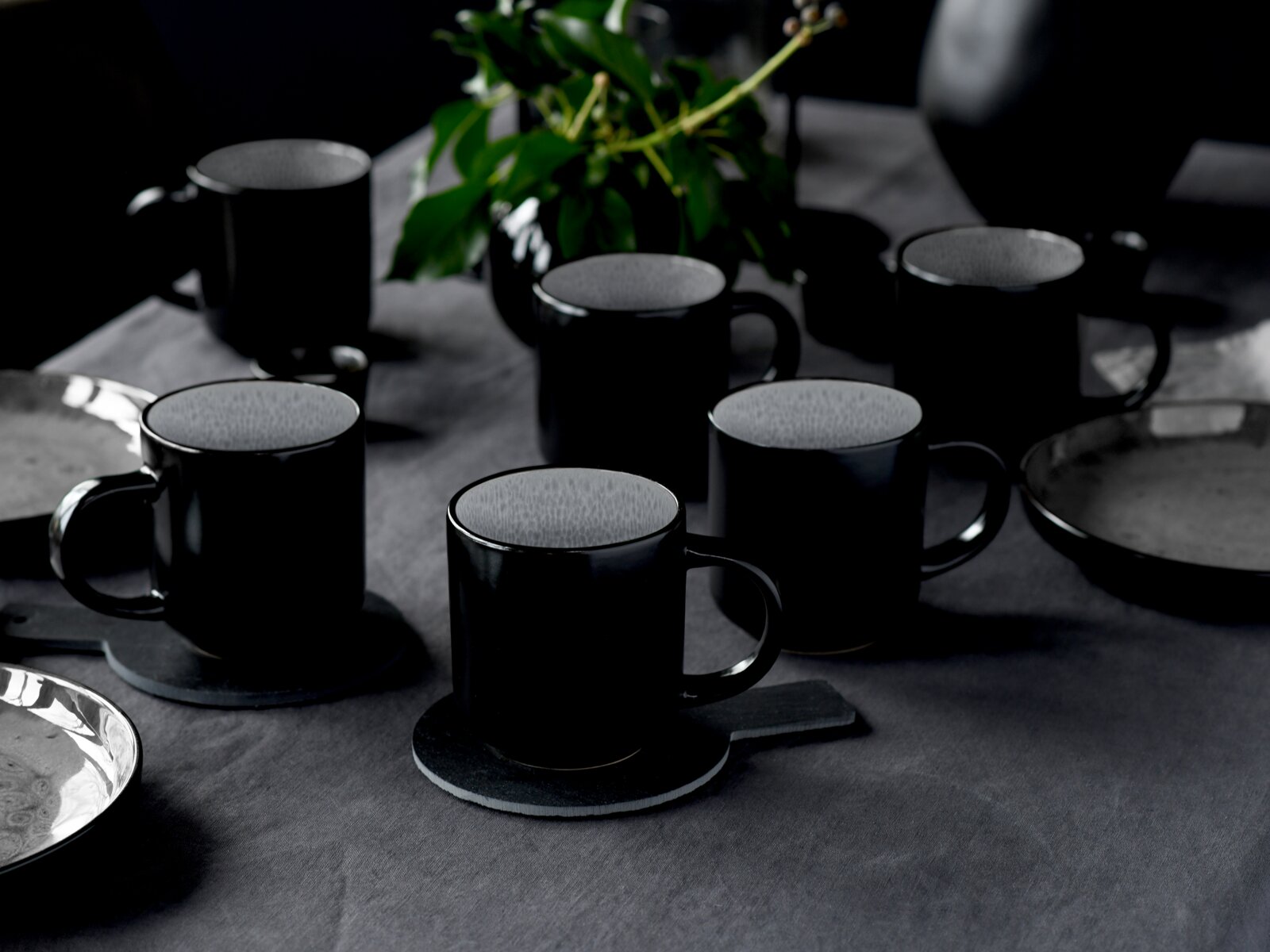 Mehrere schwarze Keramik-Kaffeetassen mit hell gesprenkelter Innenseite auf einem dunkel gedeckten Tisch, teils auf Untersetzern, fotografiert aus leicht erhöhter schräger Perspektive (3/4-Ansicht)