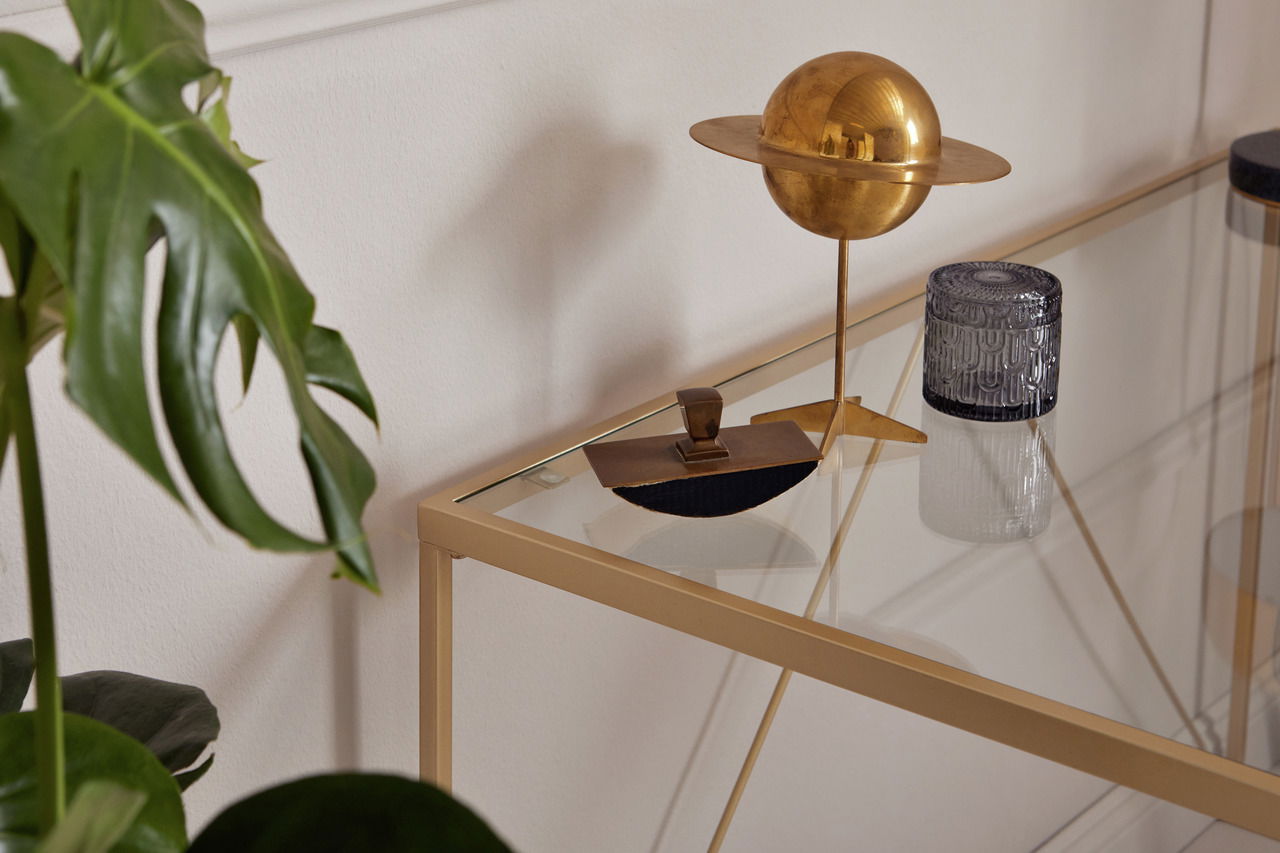 Nahaufnahme eines eleganten Schreibtisches mit Glasplatte und goldfarbenem Rahmen, dekoriert mit einer goldenen Skulptur und einer Pflanze im Hintergrund.