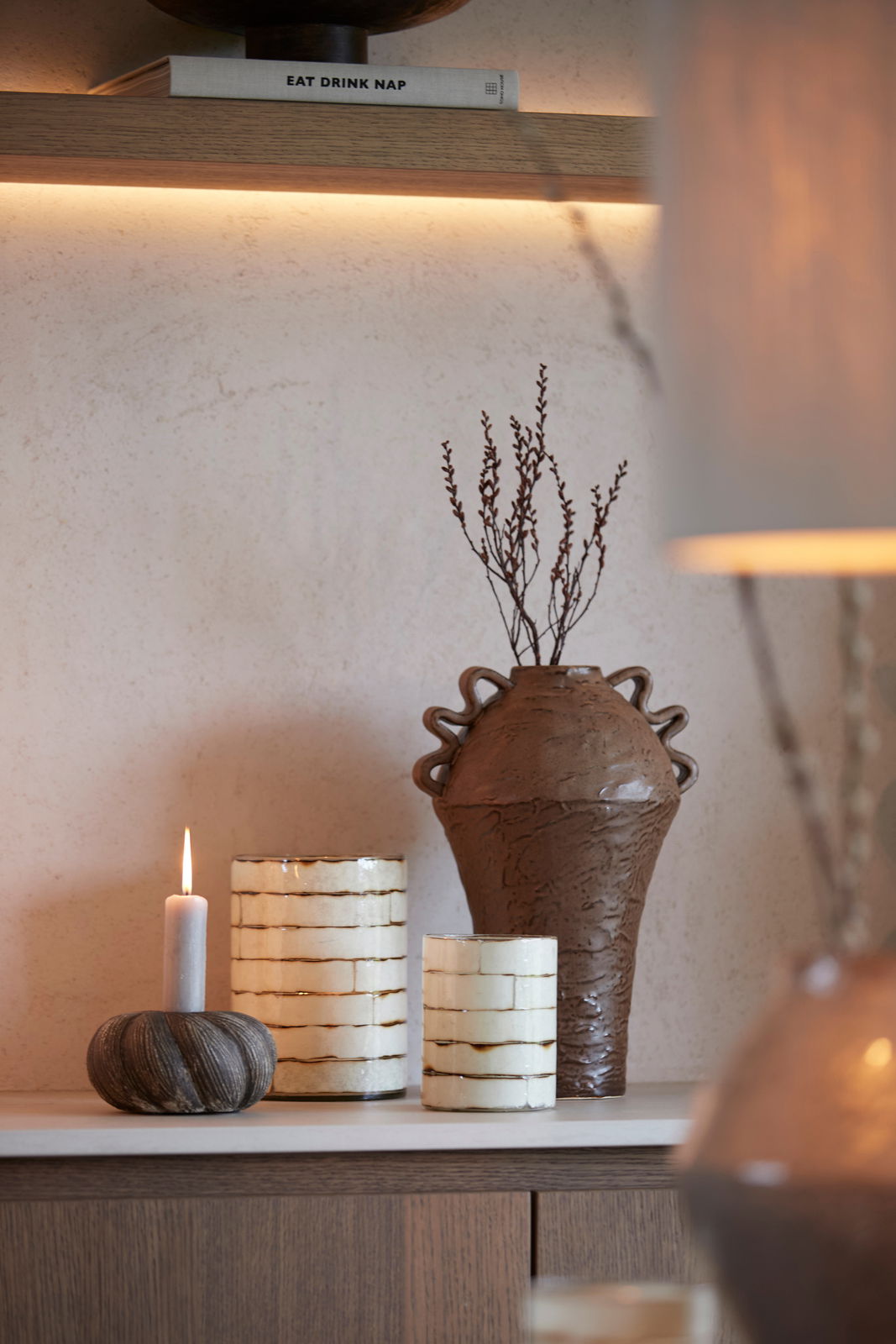 Light&Living Teelicht DRONERI 20cm Stimmungsvolle Szene mit brennendem Teelicht in einem rustikalen Kerzenhalter, umgeben von dekorativen Vasen und Zweigen, seitliche Perspektive.