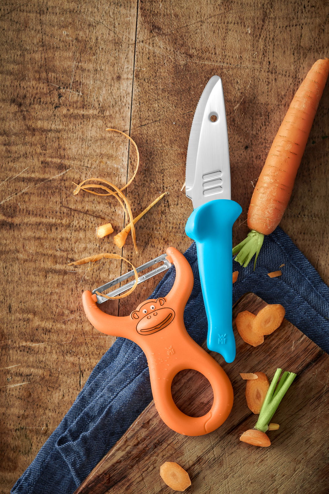 Koch-Set bestehend aus einem blauen Kindermesser und einem orangefarbenen Sparschäler mit Affenmotiv, auf einem Holztisch mit Karottenstücken, aus der Vogelperspektive.
