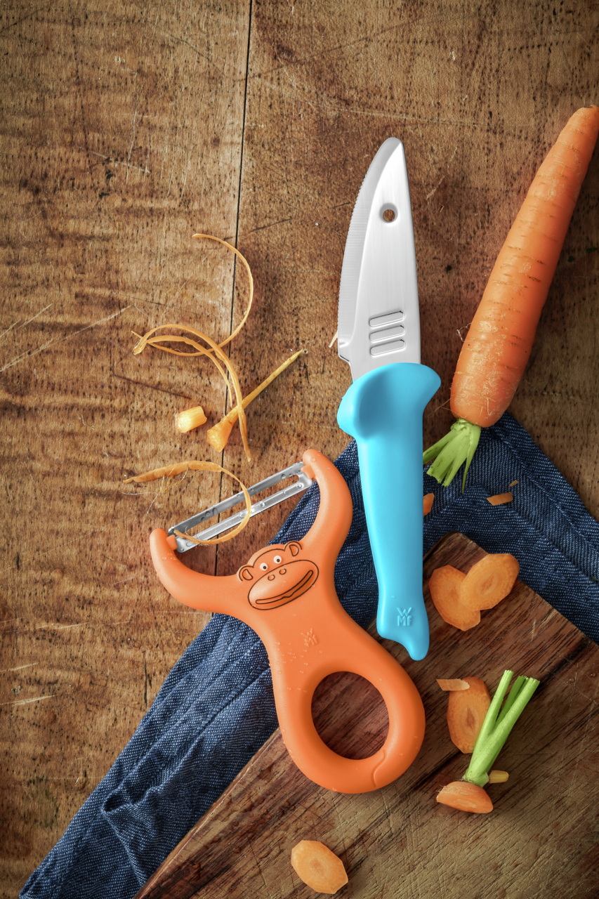 Koch-Set bestehend aus einem blauen Kindermesser und einem orangefarbenen Sparschäler mit Affenmotiv, auf einem Holztisch mit Karottenstücken, aus der Vogelperspektive.