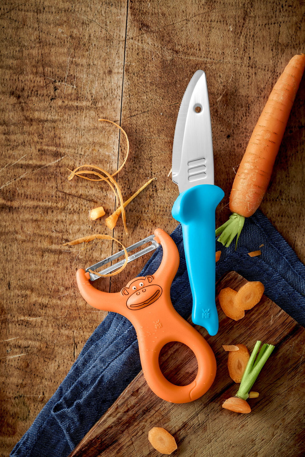 WMF Kinderkoch-Set 2-tlg. WILLY MIA FRED Koch-Set bestehend aus einem blauen Kindermesser und einem orangefarbenen Sparschäler mit Affenmotiv, auf einem Holztisch mit Karottenstücken, aus der Vogelperspektive.