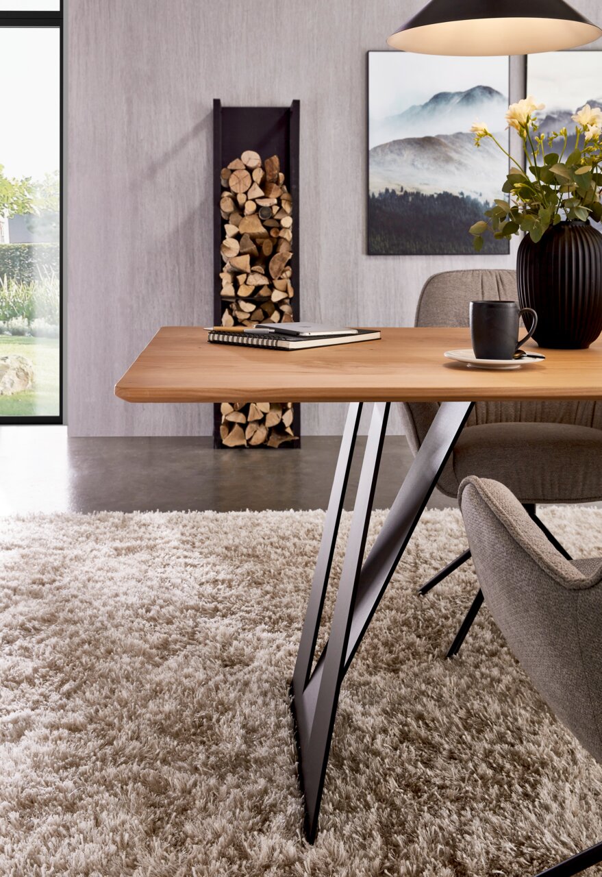 Moderne eettafel met een lichtgekleurd houten blad en zwarte, V-vormige metalen poten in de woon-/eetkamer, afgebeeld in zijaanzicht op de tafelrand en het onderstel.