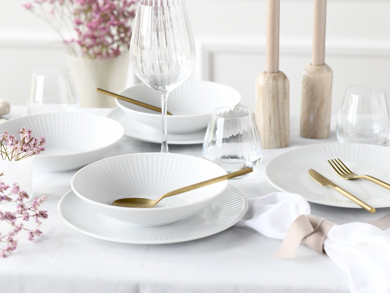 Heller, elegant gedeckter Tisch mit weißen, geriffelten Porzellantellern und Schalen, goldfarbenem Besteck sowie Wein- und Wassergläsern; Perspektive: leicht erhöht und frontal aus Tischhöhe.