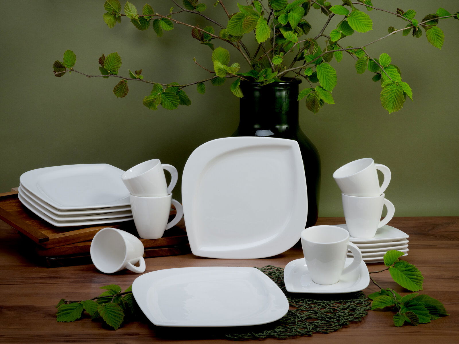 Creatable Kaffeeservice 18-tlg. MELBOURNE Kaffeeservice 18-teilig, bestehend aus weißen Tassen und Tellern, arrangiert auf einem Holztisch mit grünen Blättern im Hintergrund, seitliche Perspektive.
