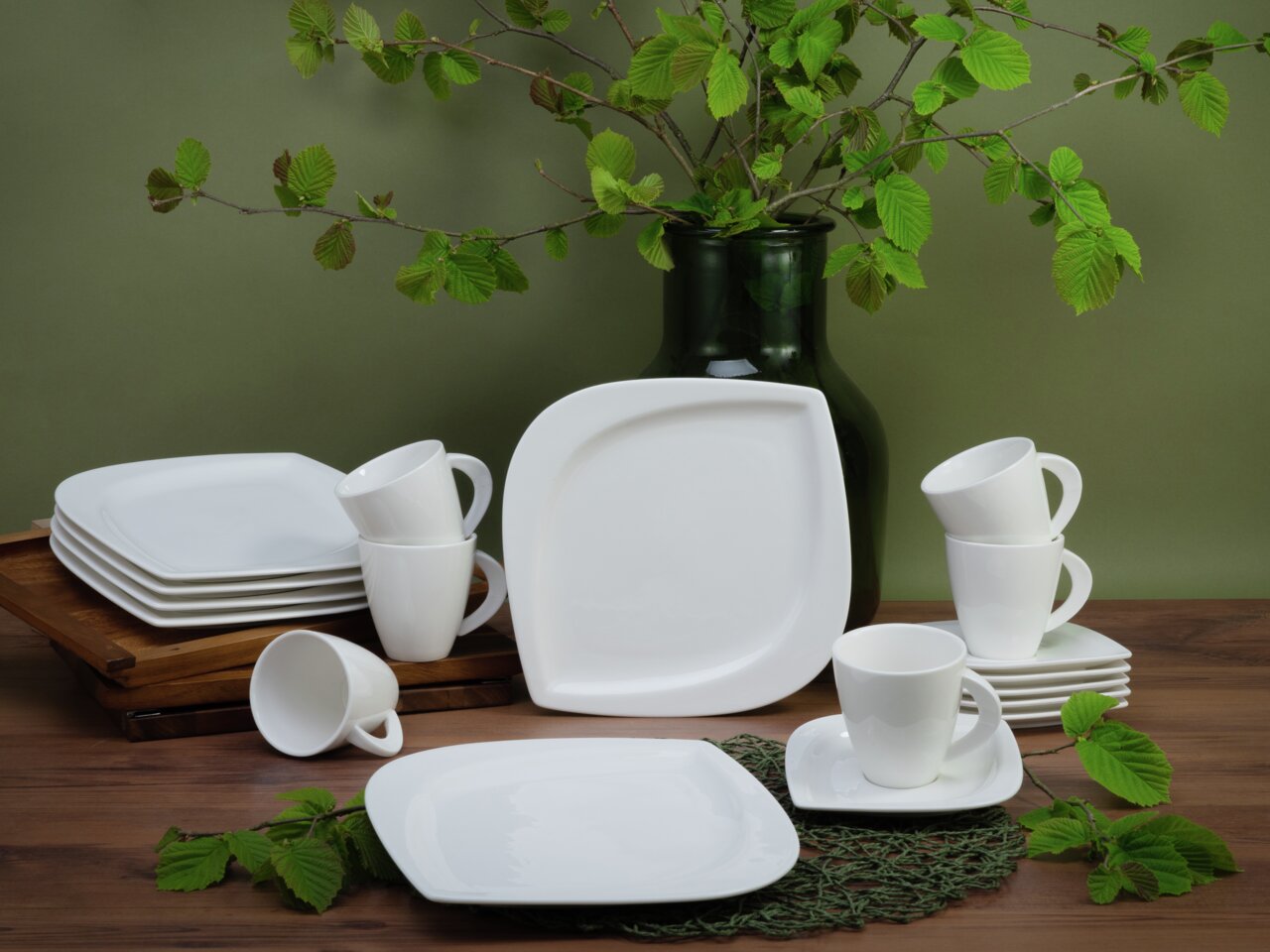 Creatable Kaffeeservice 18-tlg. MELBOURNE Kaffeeservice 18-teilig, bestehend aus weißen Tassen und Tellern, arrangiert auf einem Holztisch mit grünen Blättern im Hintergrund, seitliche Perspektive.