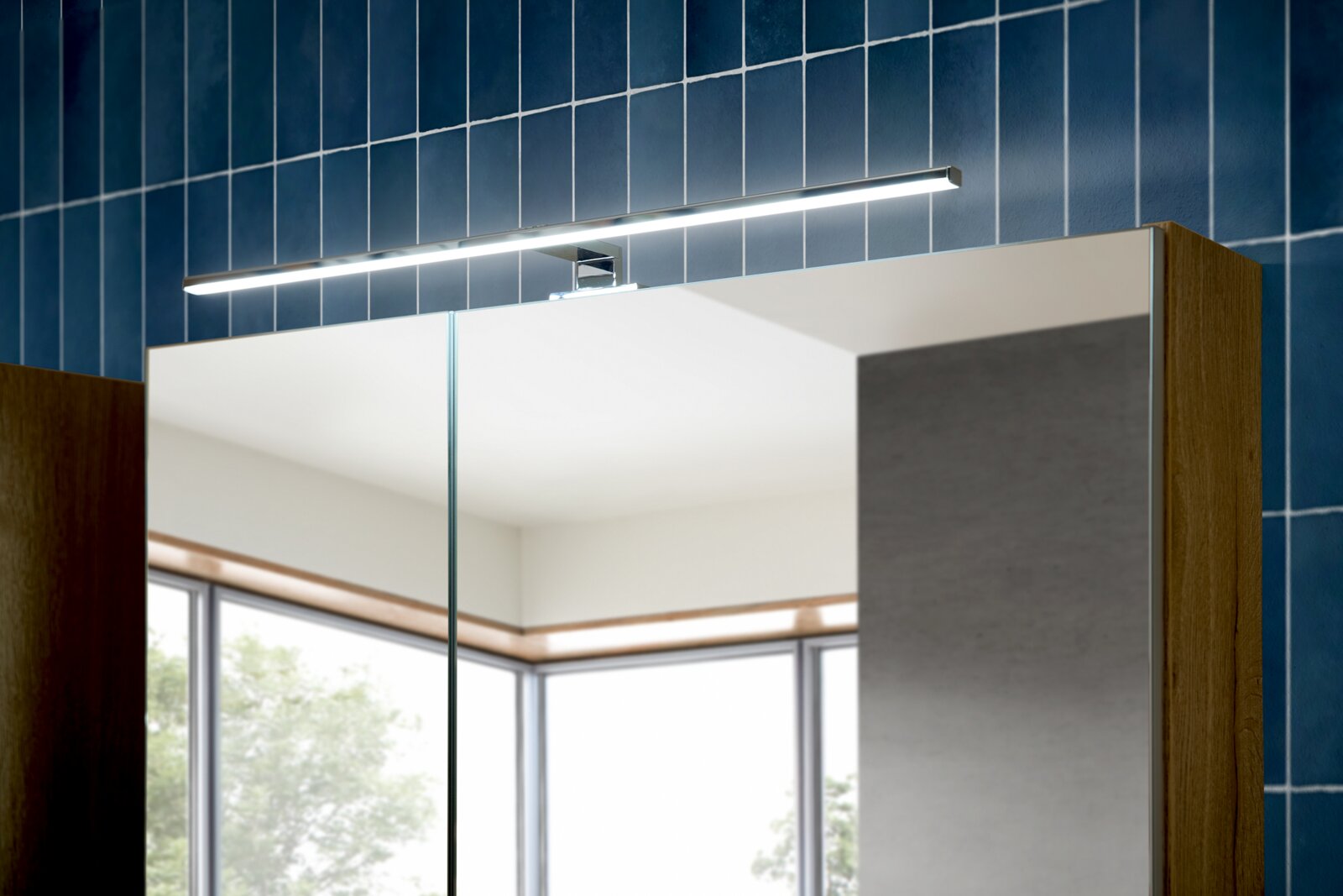 LED-Spiegelleuchte in Chrom über einem Badezimmerspiegelschrank mit Holzseiten vor blauer Fliesenwand, frontal leicht von rechts oben aufgenommen