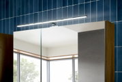LED-Spiegelleuchte in Chrom über einem Badezimmerspiegelschrank mit Holzseiten vor blauer Fliesenwand, frontal leicht von rechts oben aufgenommen