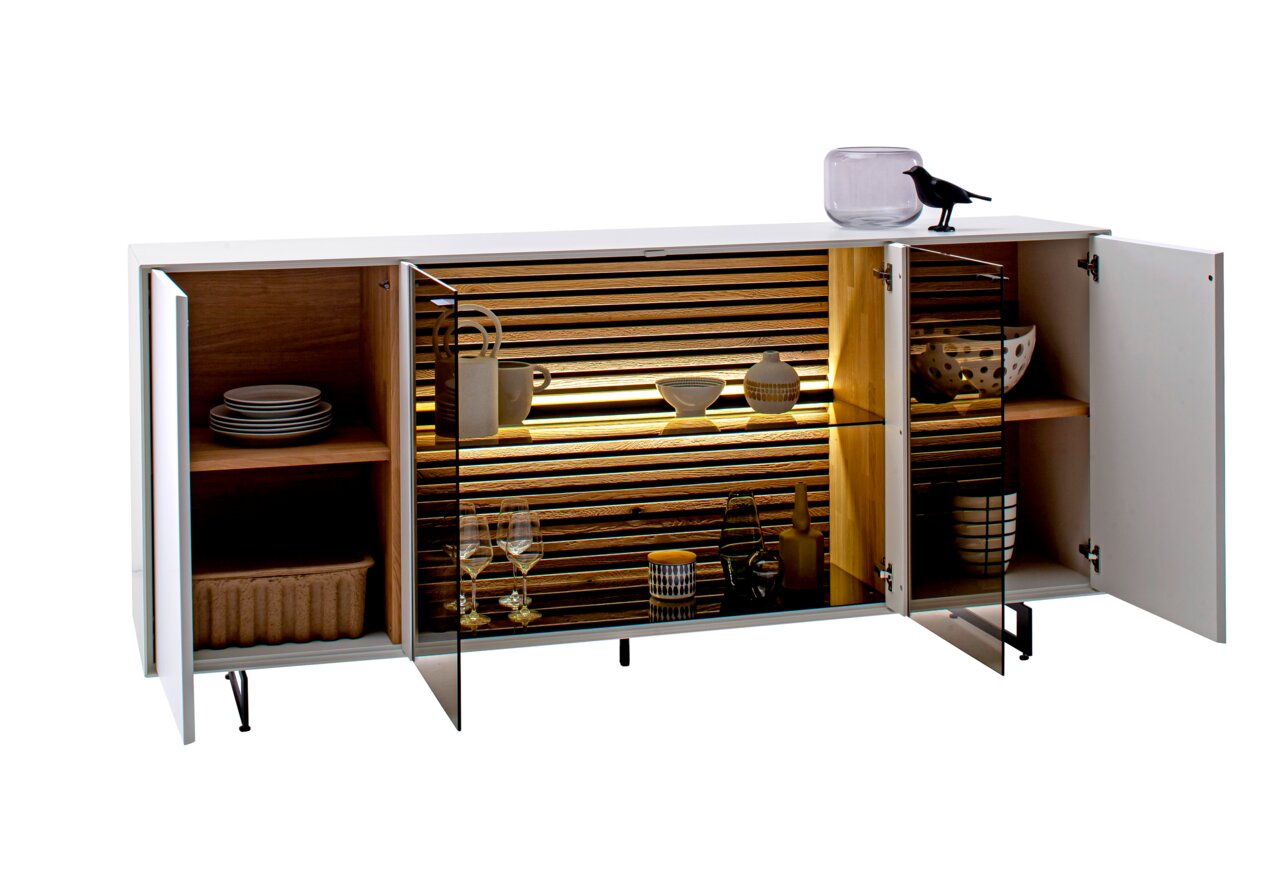 Modernes Sideboard mit offenen Türen, das von vorne gezeigt wird. Es hat eine weiße Außenseite, Holzelemente im Inneren und integrierte Beleuchtung. Dekorative Gegenstände wie Geschirr und Gläser sind sichtbar.