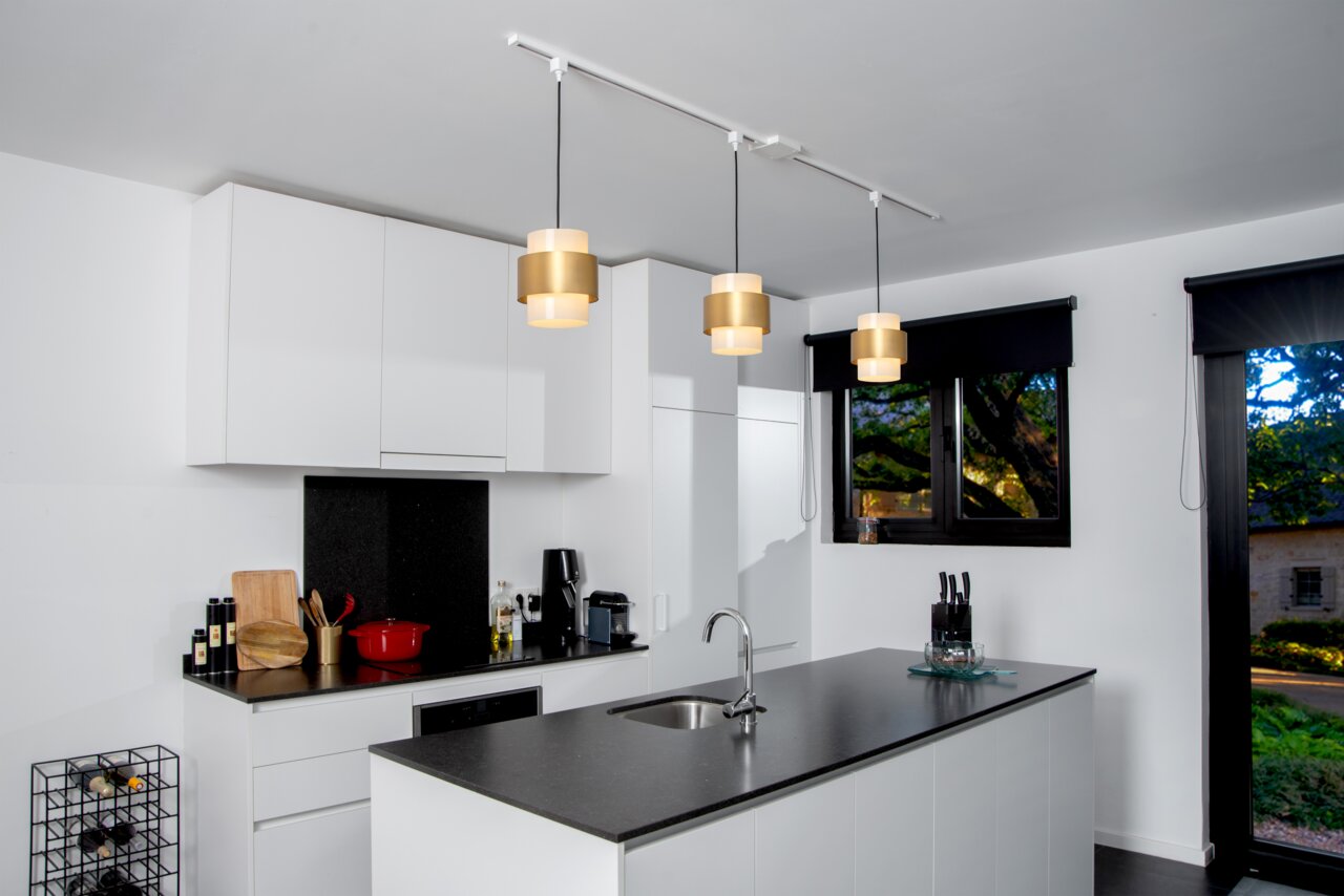 Moderne Küche mit drei goldenen Pendelleuchten, die an einem weißen Adapter von der Decke hängen. Perspektive von der Seite der Kücheninsel.