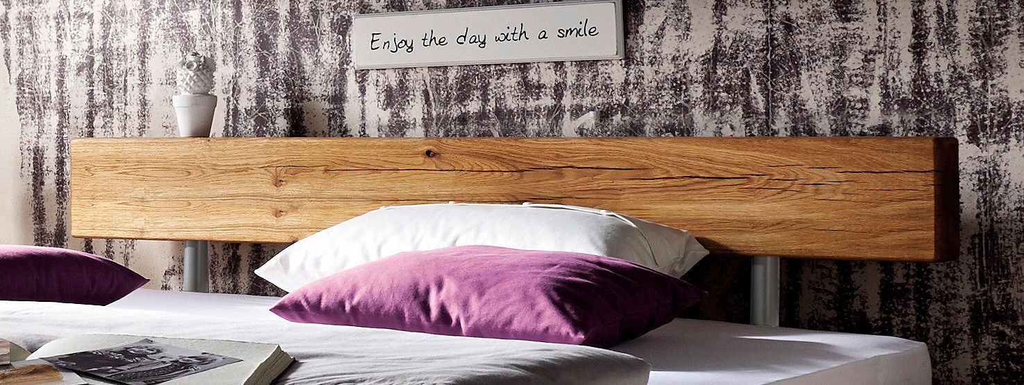 Nahaufnahme eines Balkenbetts mit rustikalem Holzkopfteil, dekoriert mit lila und weißen Kissen, im Vordergrund. Im Hintergrund ein Schild mit der Aufschrift 'Enjoy the day with a smile'.
