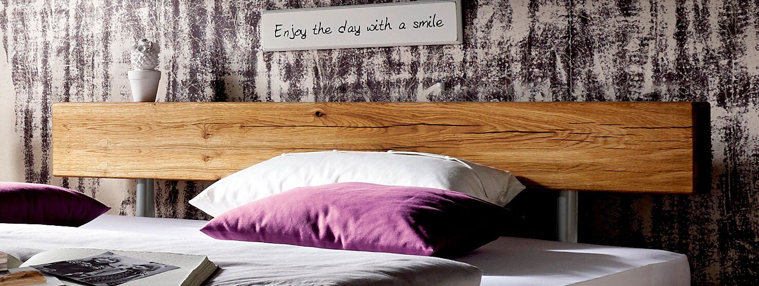 Nahaufnahme eines Balkenbetts mit rustikalem Holzkopfteil, dekoriert mit lila und weißen Kissen, im Vordergrund. Im Hintergrund ein Schild mit der Aufschrift 'Enjoy the day with a smile'.